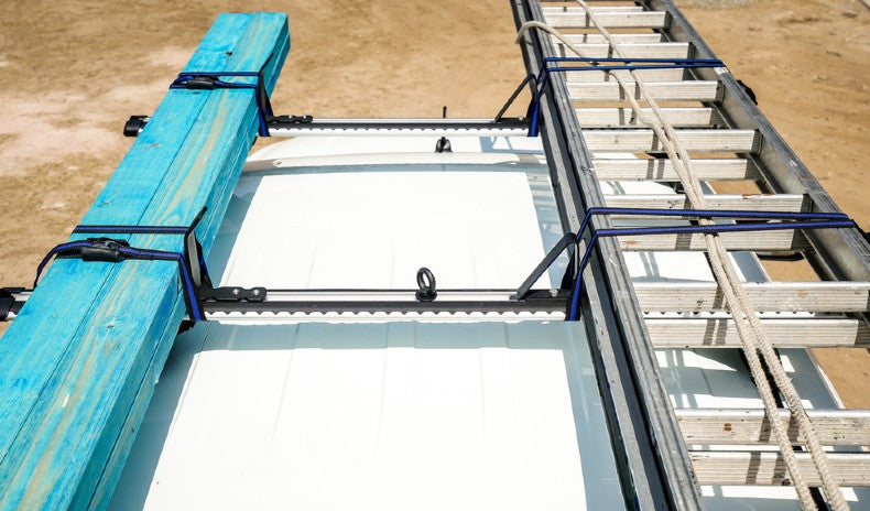 A commercial van roof fitted with a ladder rack carrying timber and a ladder, secured with load stops and straps